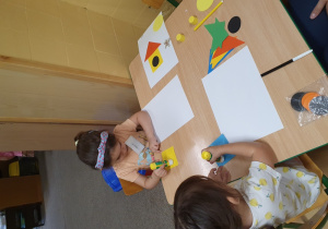 Dzieci przyklejają papierowe figury geometryczne na kartkę tworząc budę.