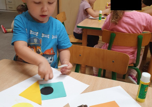 Chłopiec przykleja papierowe figury geometryczne na kartkę tworząc budę.