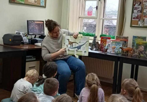 Pani Bibliotekarka pokazuje dzieciom książkę z ilustracjami dinozaurów