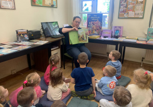 Dzieci siedzą na poduszkach i słuchają pani bibliotekarki.