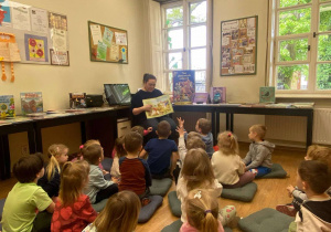 Dzieci siedzą na poduszkach i słuchają pani bibliotekarki.