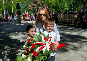 Dzieci wraz z nauczycielką trzymają biało czerwone kwiaty.